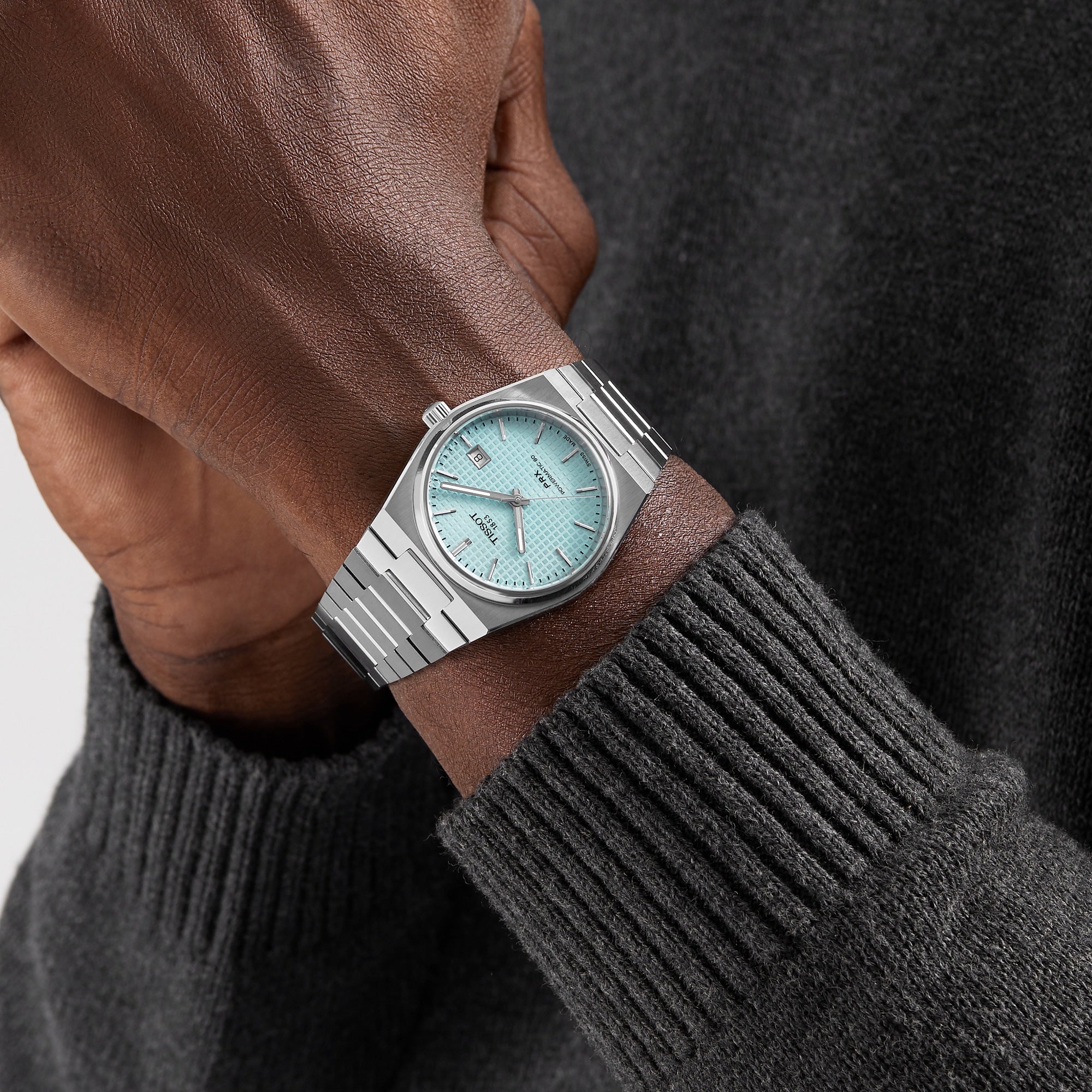 A person wearing the Tissot PRX Powermatic 80 with a stainless steel case, light blue dial, and silver band, dressed in a dark gray sweater.