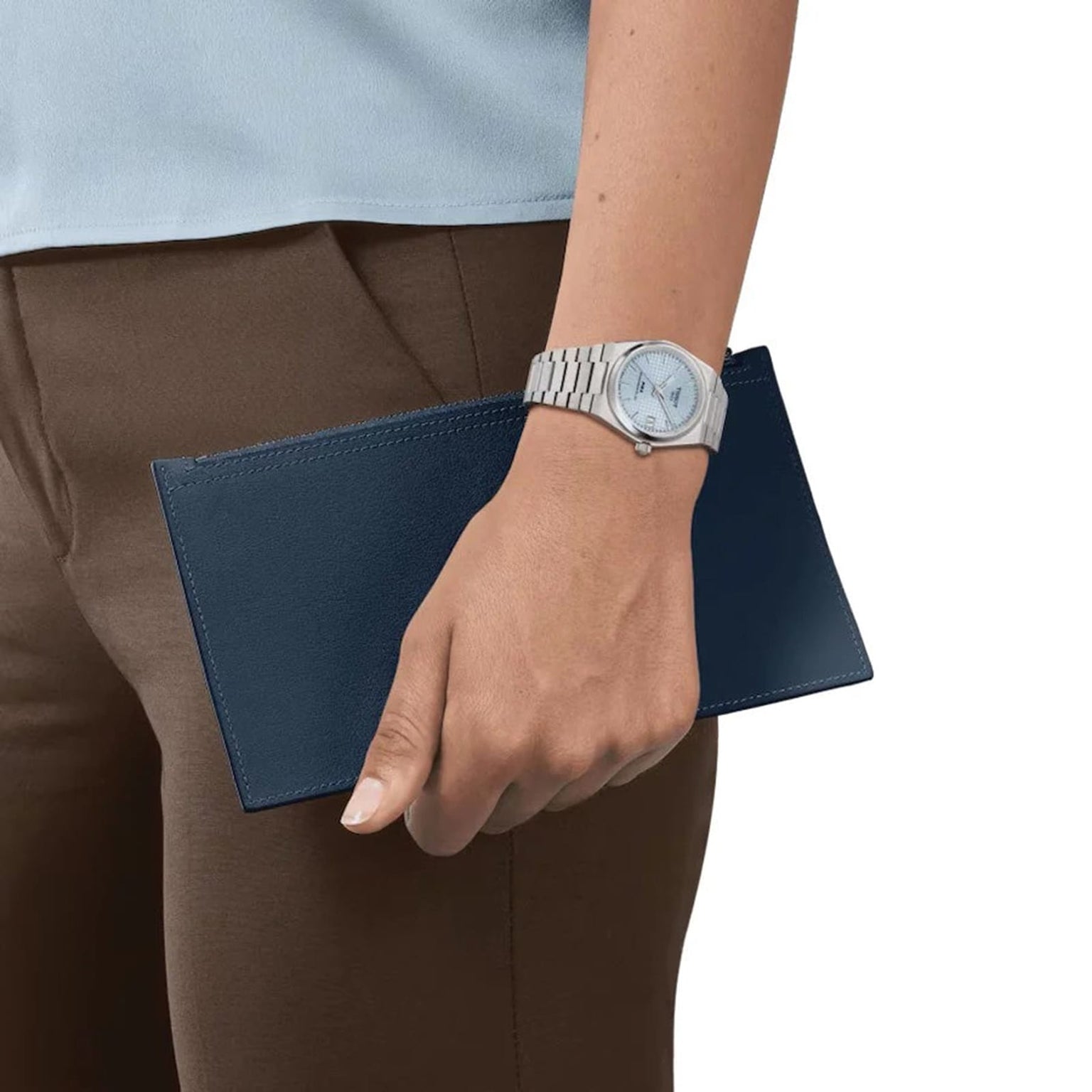 Someone wearing a Tissot PRX Powermatic 80 with an ice blue dial holds a dark blue rectangular wallet or clutch against their brown pants.