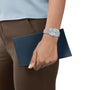 Someone wearing a Tissot PRX Powermatic 80 with an ice blue dial holds a dark blue rectangular wallet or clutch against their brown pants.