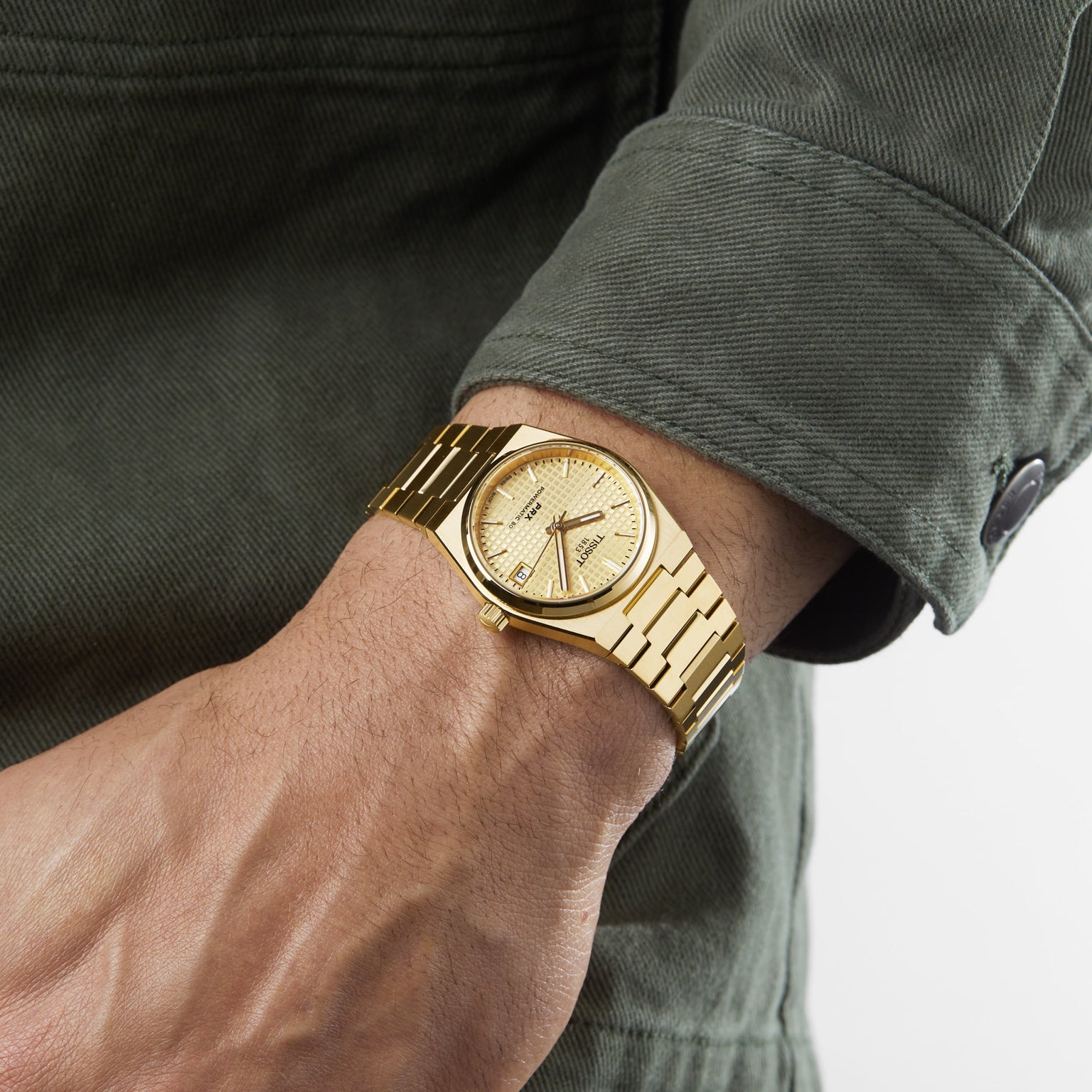 Wearing a Tissot PRX Powermatic 80 with a gold-PVD case and waffle dial, the person pairs it with a green jacket featuring buttoned cuffs.