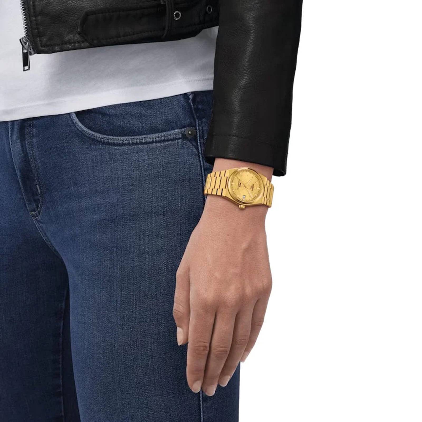 Wearing blue jeans, a white shirt, and a black leather jacket, someone displays a Tissot PRX Powermatic 80 with a gold-PVD case and waffle dial pattern on their left wrist.