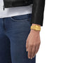 Wearing blue jeans, a white shirt, and a black leather jacket, someone displays a Tissot PRX Powermatic 80 with a gold-PVD case and waffle dial pattern on their left wrist.