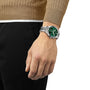 A person in a brown sweater and black pants showcases a Tissot Chrono XL Classic with a green dial and large case on their left wrist.