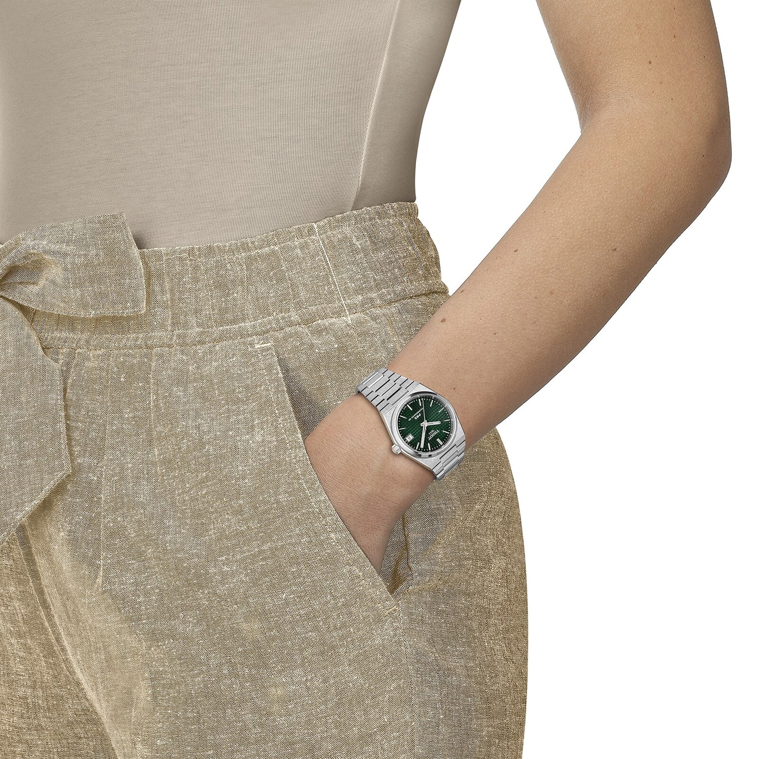 A person in beige pants and a beige top has their left hand in their pocket, showing off a Tissot PRX Powermatic 80 wristwatch with a striking green dial.