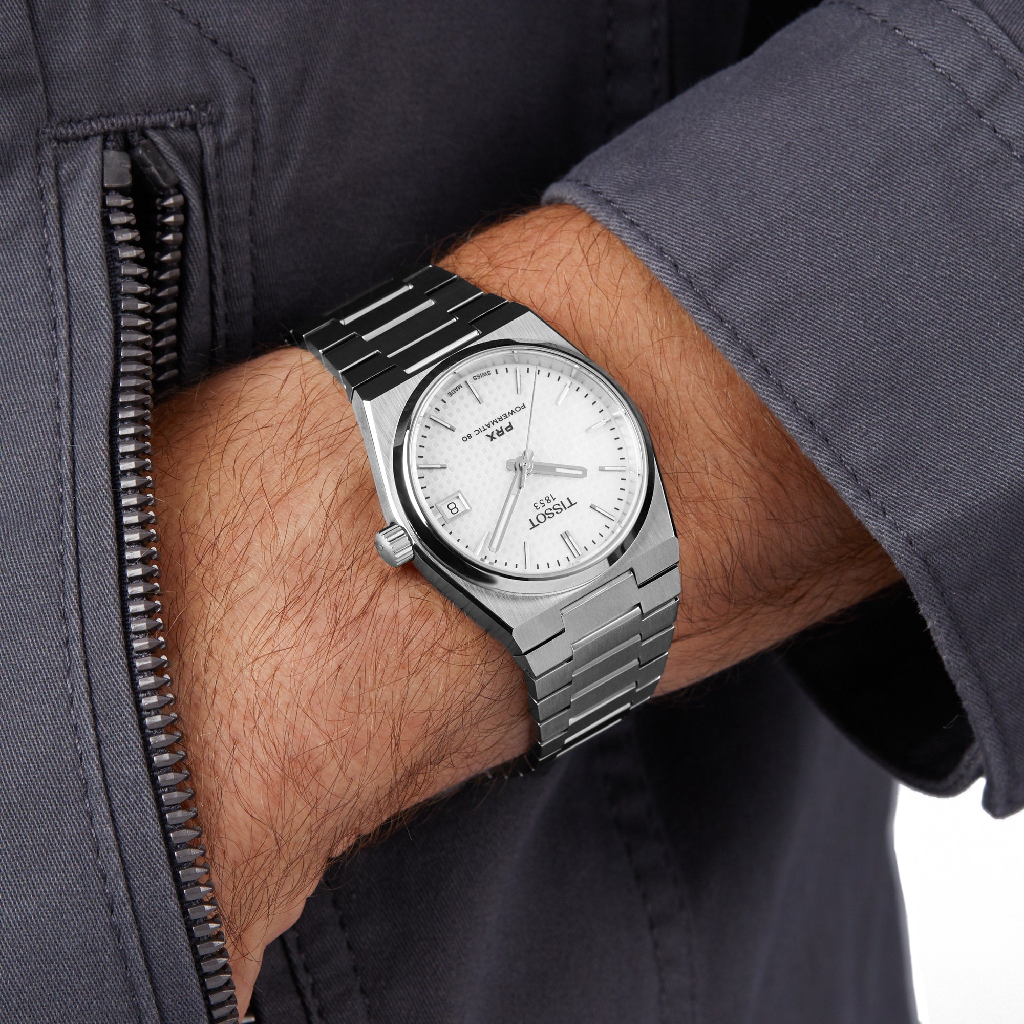 A person in a grey jacket wears a Tissot PRX Powermatic 80 with a 35mm white dial, black hour markers, and date display.