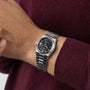 A person wearing a Tissot PRX Powermatic 80 with a silver metal bracelet and black waffle-pattern dial on their left wrist over a maroon long-sleeve shirt.