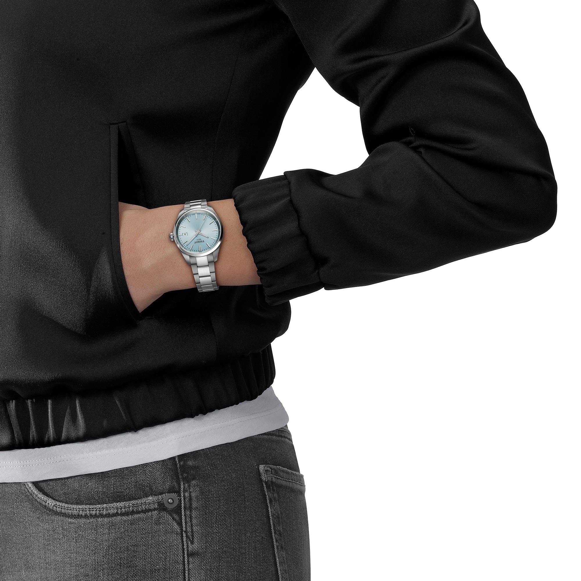 A person wearing a black jacket, gray pants, and a Tissot PR 100 silver wristwatch with a light blue face and Swiss Quartz movement on their left wrist.