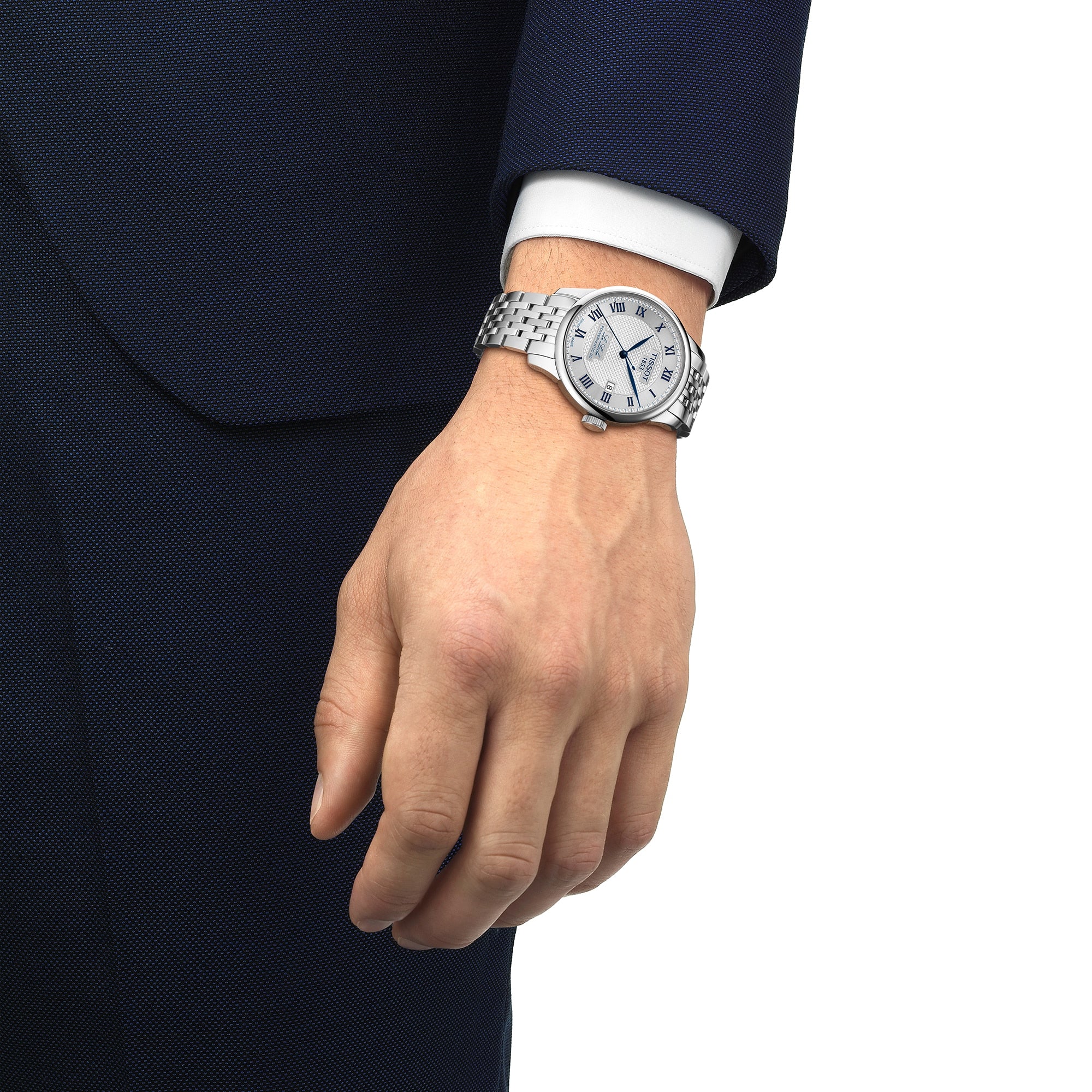 A person in a dark blue suit and white shirt wears a Tissot Le Locle Powermatic 80 20th Anniversary wristwatch with a white dial, Roman numerals, and automatic movement.