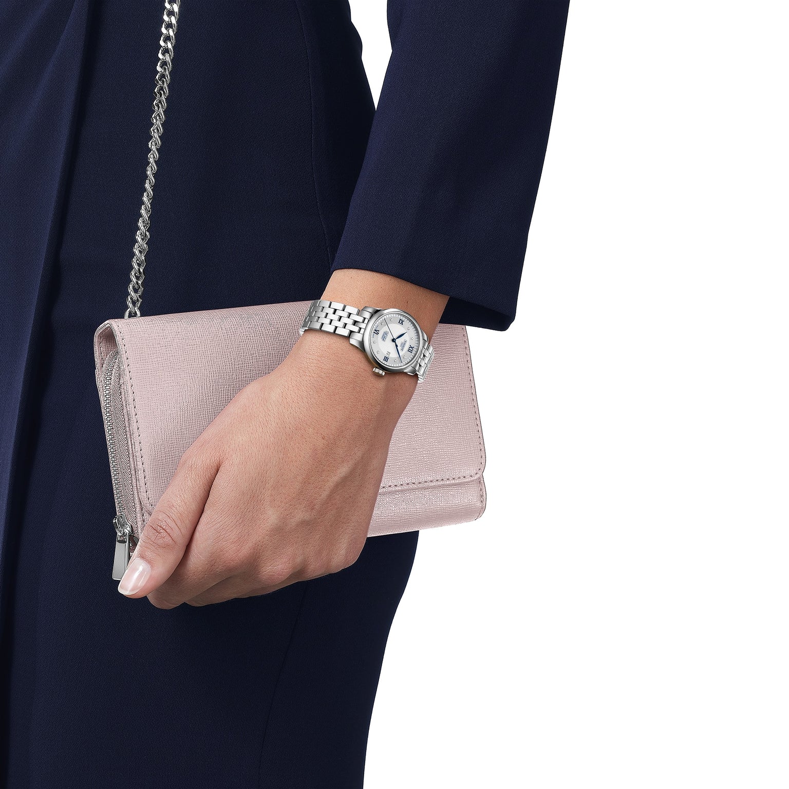 A person in a navy blue outfit holds a light pink clutch purse and wears the Tissot Le Locle Automatic 20th Anniversary wristwatch.