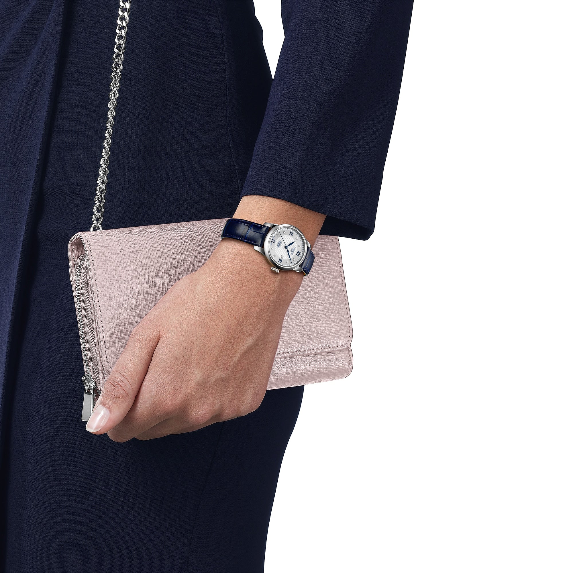 A person in a dark suit holds a light pink clutch bag, wearing the Tissot Le Locle Automatic 20th Anniversary watch with a black leather strap on their wrist.
