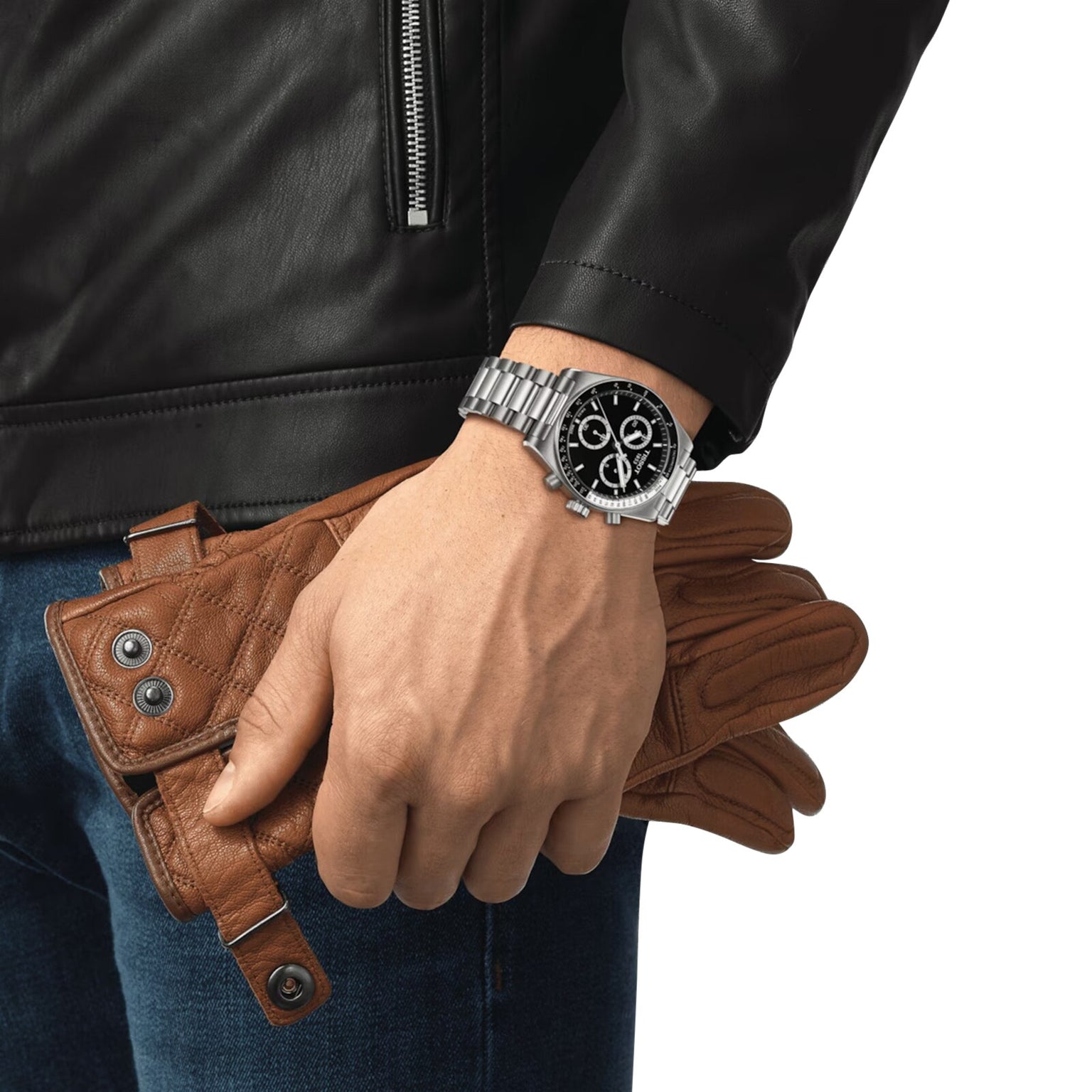 A person in a black leather jacket and jeans holds brown leather gloves and wears a silver Tissot PR516 Chronograph watch.