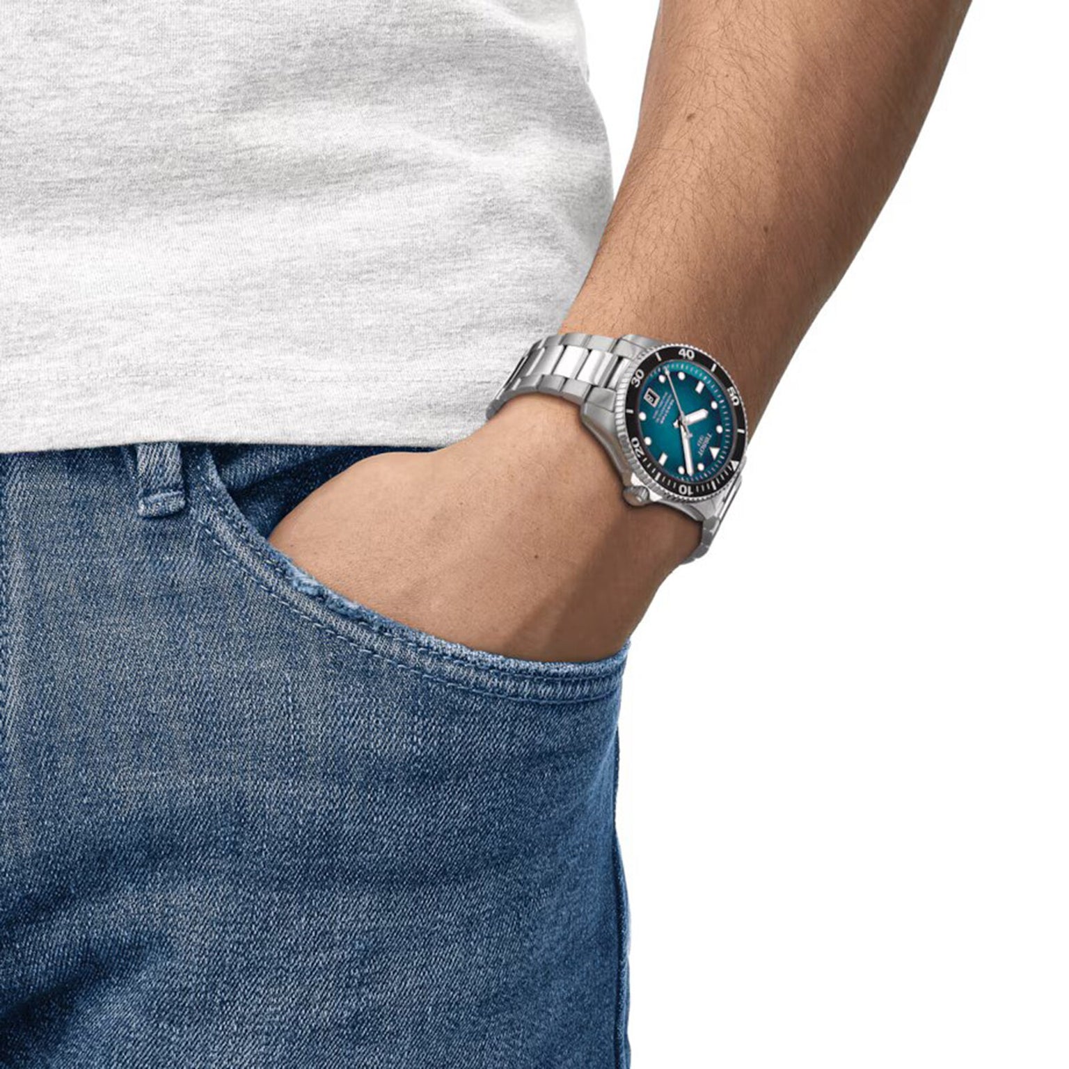 A person wearing a Tissot Seastar 1000 Powermatic 80 with Super-LumiNova® and a blue dial has their hand in the pocket of blue jeans, dressed in a light gray t-shirt.
