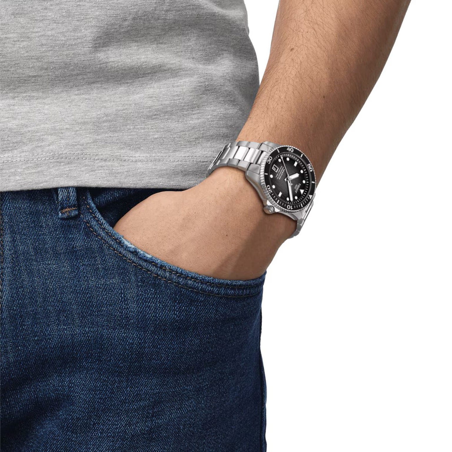A person wearing a Tissot Seastar 1000 Powermatic 80 with a black face and bezel, dressed in a grey t-shirt and blue jeans, one hand in their pocket.