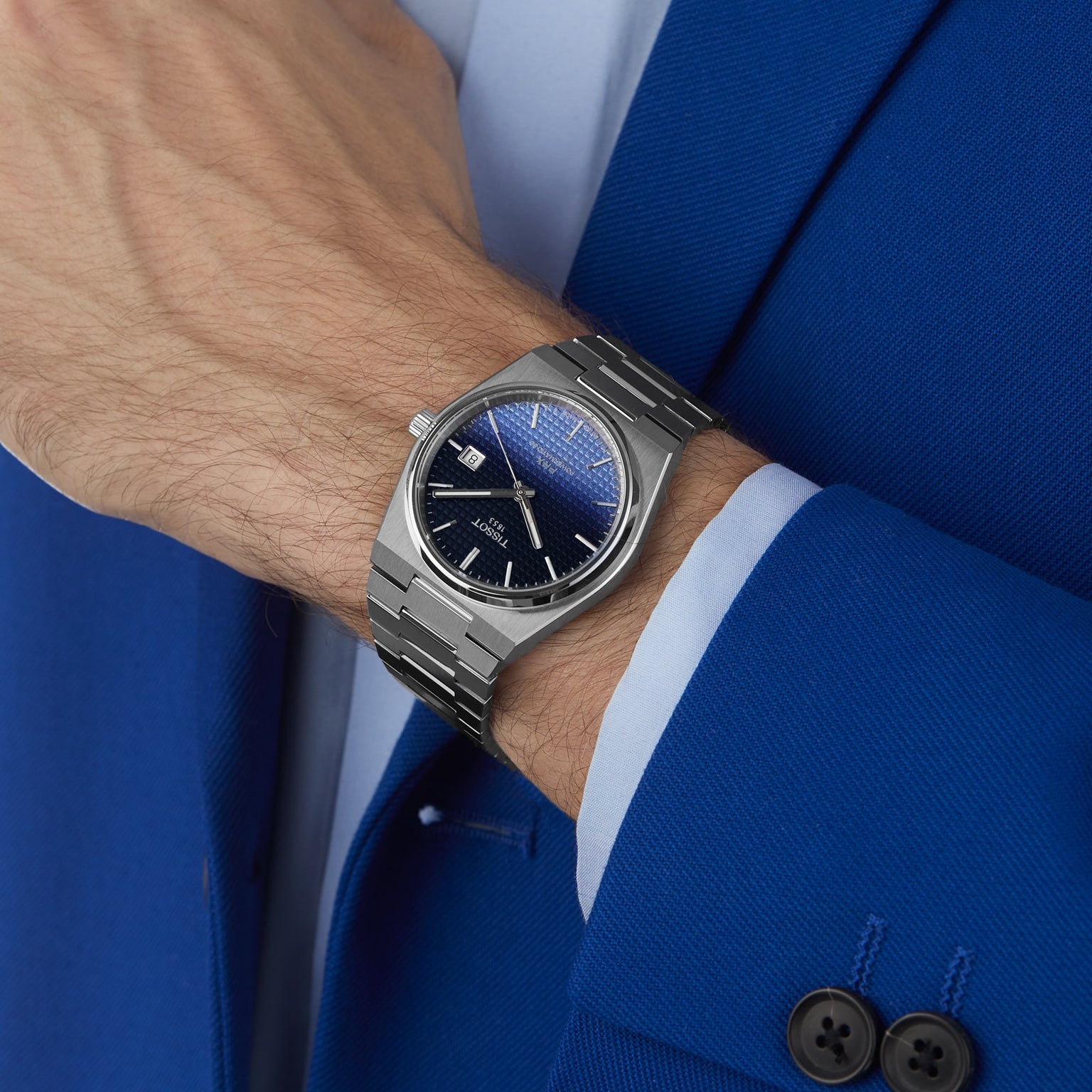 A person in a blue suit and light blue shirt wears the Tissot PRX Powermatic 80, featuring a silver case, blue textured dial, sapphire crystal, and metal bracelet on their left wrist.