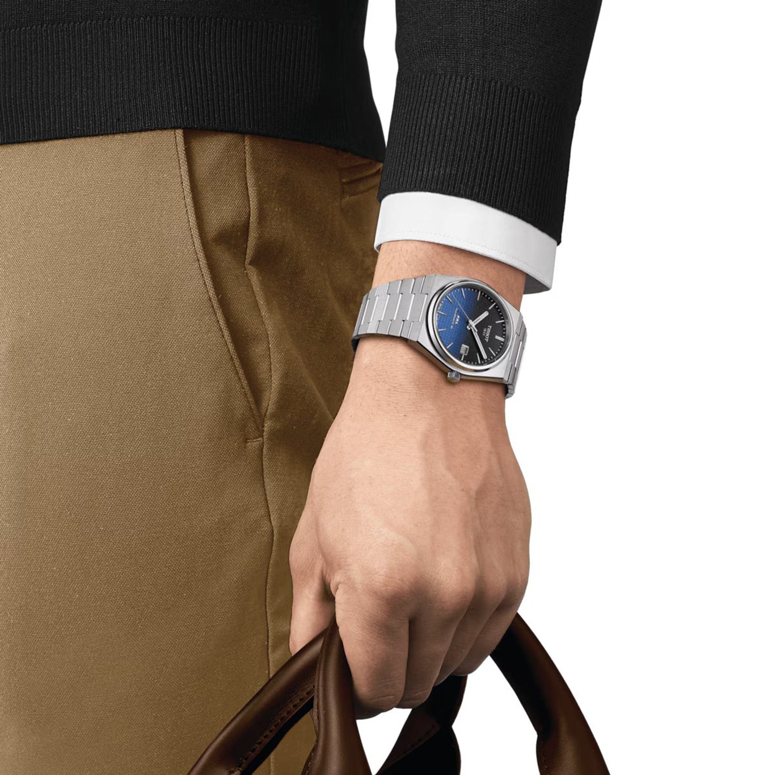 A person in a black sweater, white shirt, and tan pants holds a brown bag, showcasing a Tissot PRX Powermatic 80 with automatic movement and a striking blue face on their left wrist.