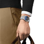 A person in a black sweater, white shirt, and tan pants holds a brown bag, showcasing a Tissot PRX Powermatic 80 with automatic movement and a striking blue face on their left wrist.