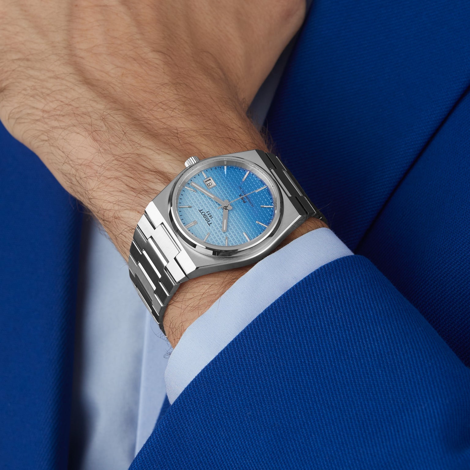 A person dressed in a blue suit jacket and light blue shirt wears a Tissot PRX Powermatic 80 wristwatch with a silver case, blue dial, sapphire crystal, and date display.