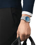 A person in a blue sweater and white shirt is holding a black object, showing a Tissot PRX Powermatic 80 wristwatch with a blue face and sapphire crystal on their left wrist.