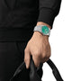 A person wearing a Tissot PRX Powermatic 80, featuring a silver case, green dial, and stainless steel bracelet, holds a black bag against a plain white background.