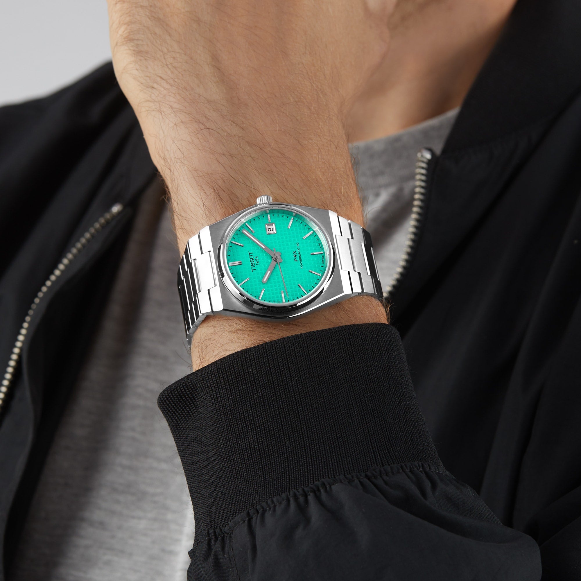 A person wearing the Tissot PRX Powermatic 80, featuring a bright turquoise dial and stainless steel bracelet, pairs it with a gray shirt and black jacket.