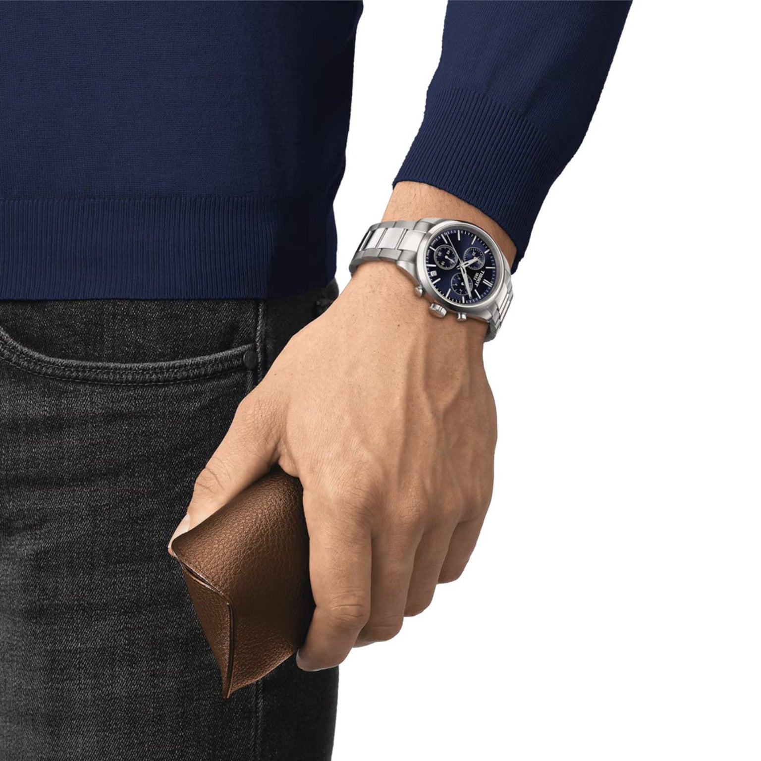 A person wearing the Tissot PR 100 Chronograph with Super-LumiNova® coating and a blue long-sleeve shirt holds a brown leather wallet.