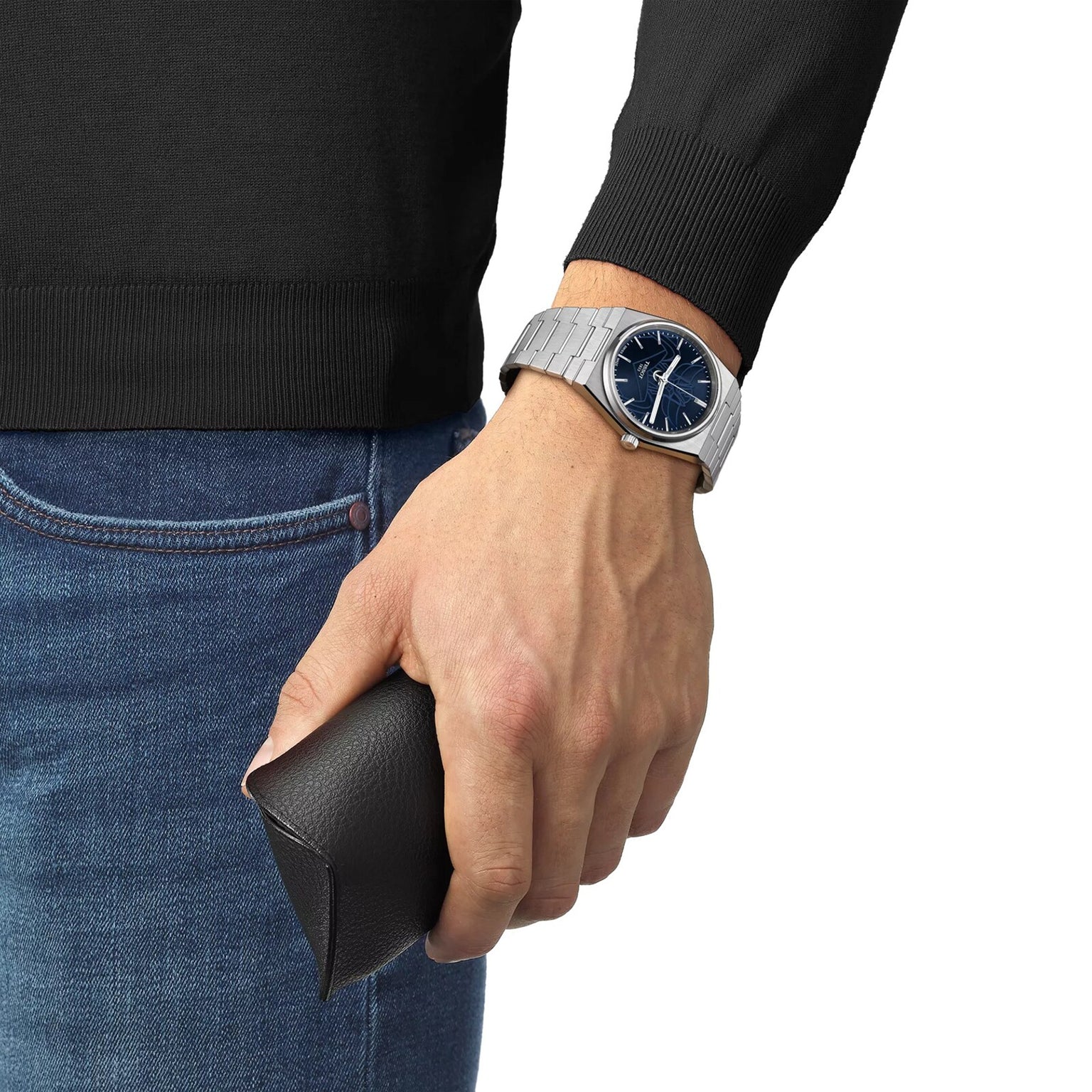 A person in jeans and a black sweater wears a Tissot PRX UFO Robot Grendizer silver watch and holds a black leather case.