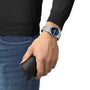 A person in jeans and a black sweater wears a Tissot PRX UFO Robot Grendizer silver watch and holds a black leather case.