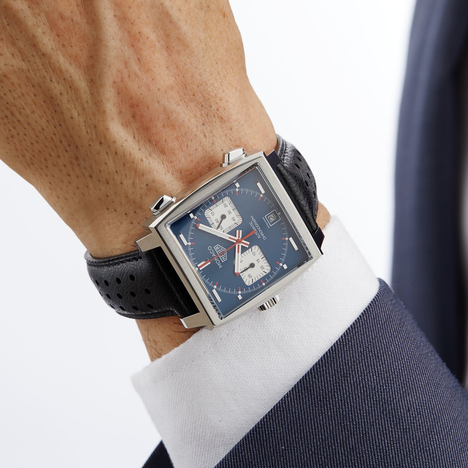 Channeling Steve McQueen style, this person wears a TAG Heuer Monaco Chronograph with a blue dial, white subdials, and black perforated leather strap, paired with a white shirt and dark suit jacket.