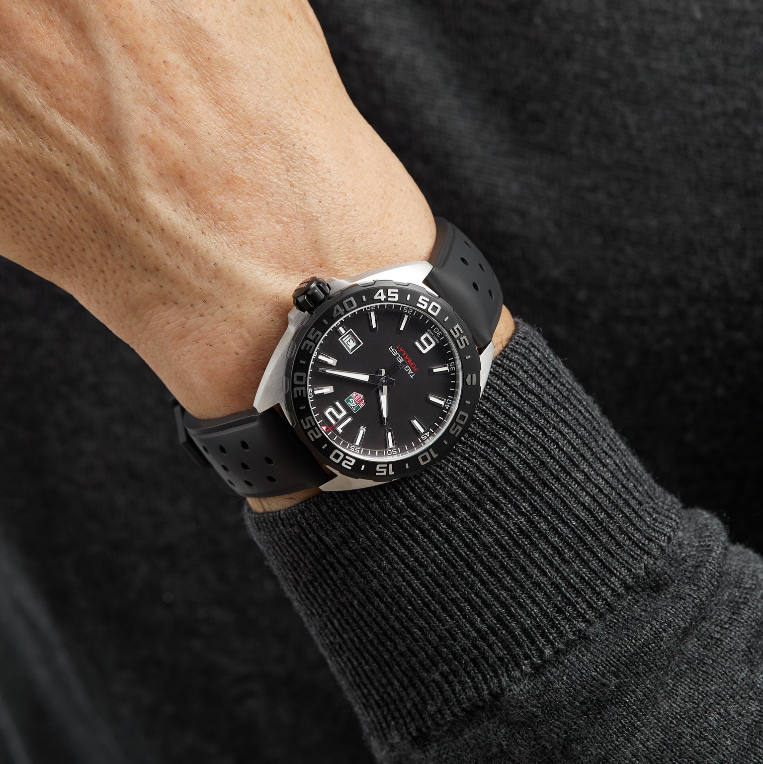 A close-up of a person wearing a TAG Heuer Formula 1 Date watch with a round dial and black perforated strap, featuring precise quartz movement, on their left wrist.