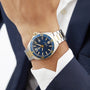 A person wearing the TAG Heuer Special Edition Formula 1 Date, featuring a blue and gold bezel with a blue dial, paired with a navy suit and white shirt.