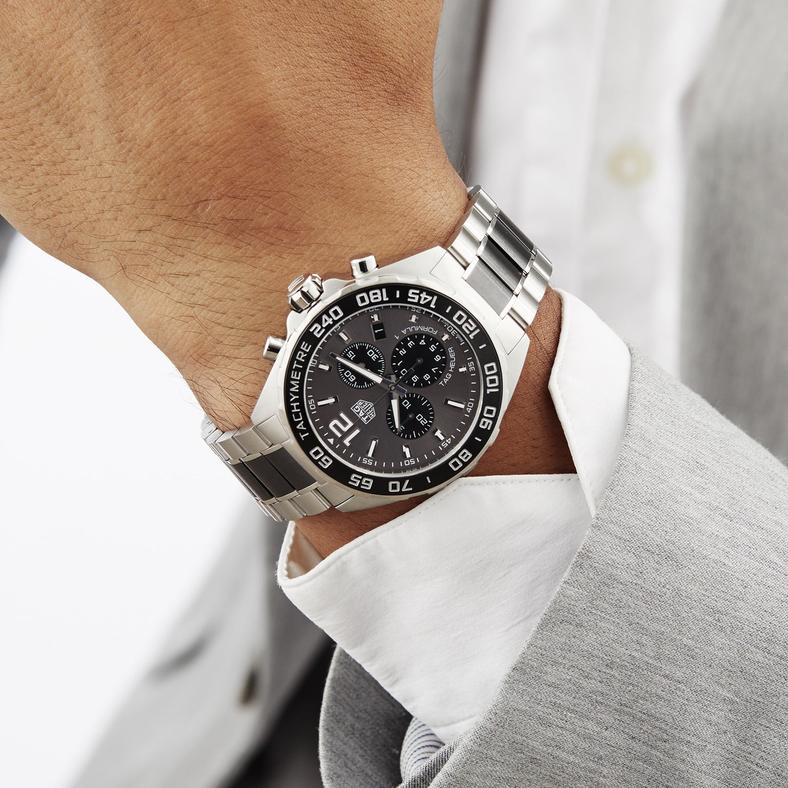 A person in a light gray suit and white shirt wears a TAG Heuer Formula 1 Chronograph, a silver wristwatch with a black dial and motor-racing inspired design.