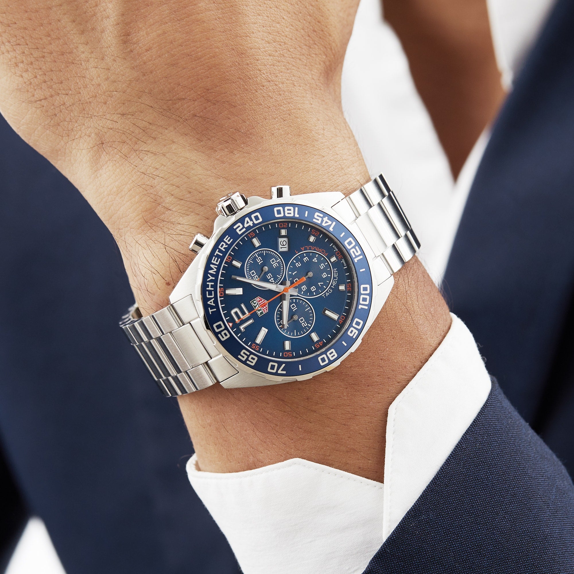A suited individual wears a TAG Heuer Formula 1 Chronograph with a stainless steel case, blue dial, and tachymeter bezel on their left wrist.