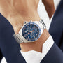 A suited individual wears a TAG Heuer Formula 1 Chronograph with a stainless steel case, blue dial, and tachymeter bezel on their left wrist.