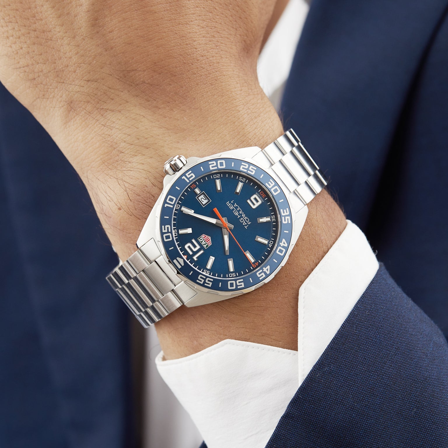 A person wearing a TAG Heuer Formula 1 Date watch with a blue sunray dial, white markers, and stainless steel band, dressed in a dark blue suit and white shirt.