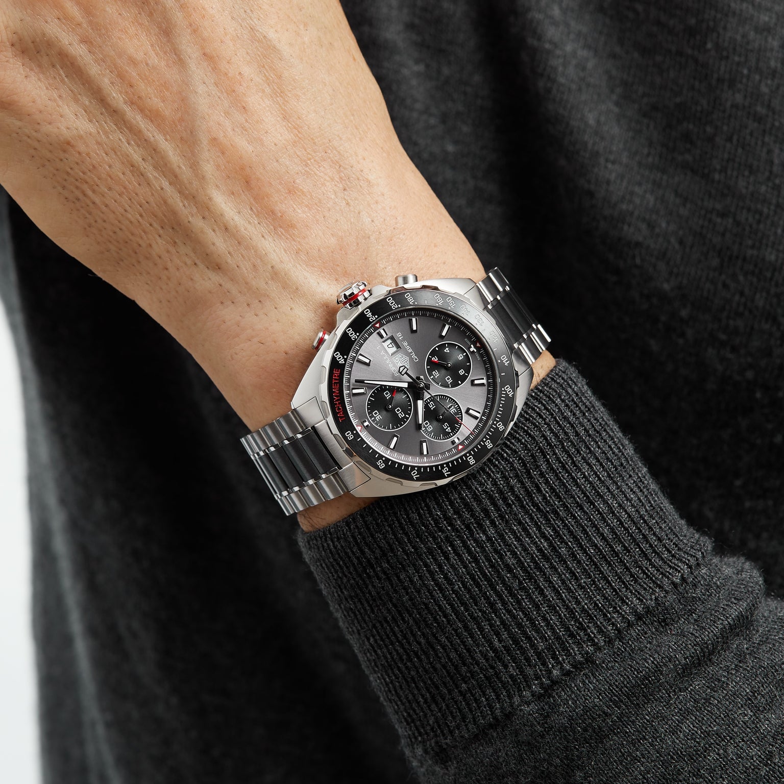 A person wearing a TAG Heuer Formula 1 Chronograph with a metal band, displayed against the sleeve of a dark sweater.