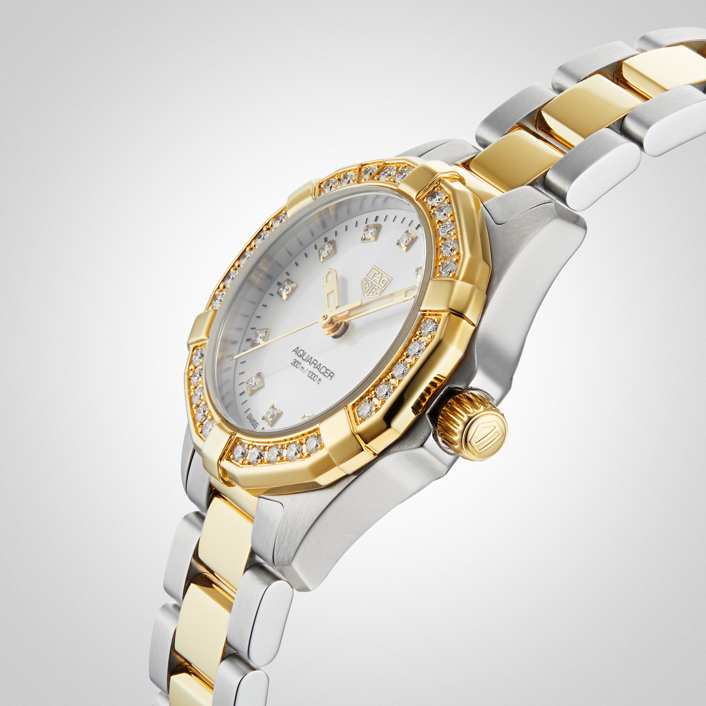 The TAG Heuer Aquaracer Date is a luxury silver and gold wristwatch with diamond accents, a white face, and a metal band, shown on a light background.
