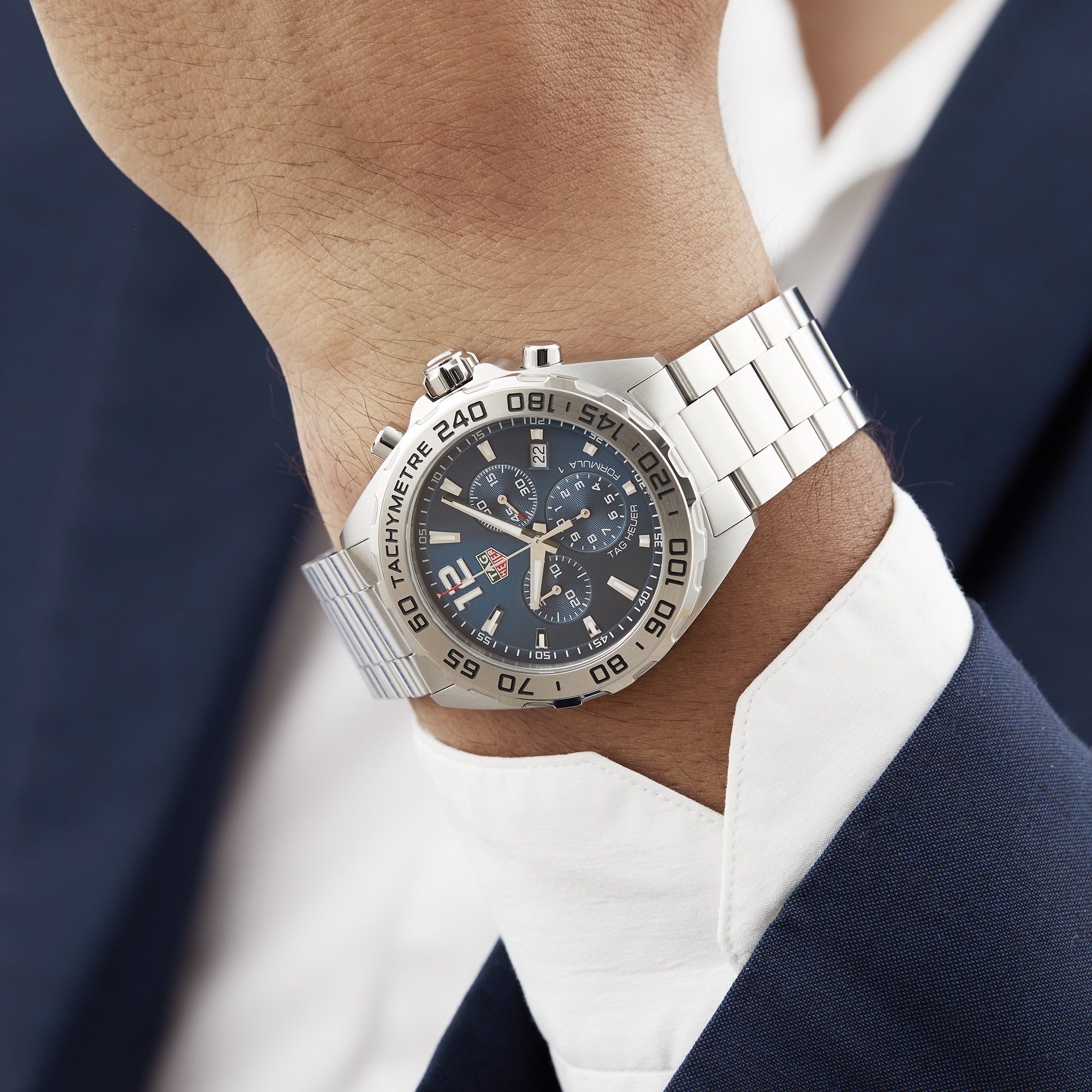 A person wearing a TAG Heuer Formula 1 Chronograph with a silver band and black dial, paired with a white shirt and dark blue suit jacket.