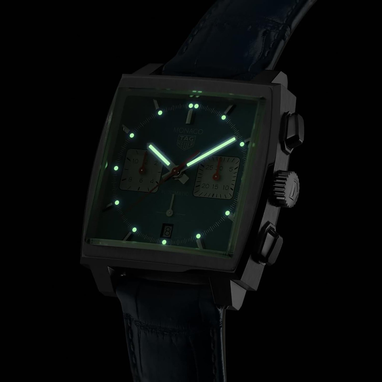 A TAG Heuer Monaco Chronograph watch with glowing green hour markers and hands in a dark setting.
