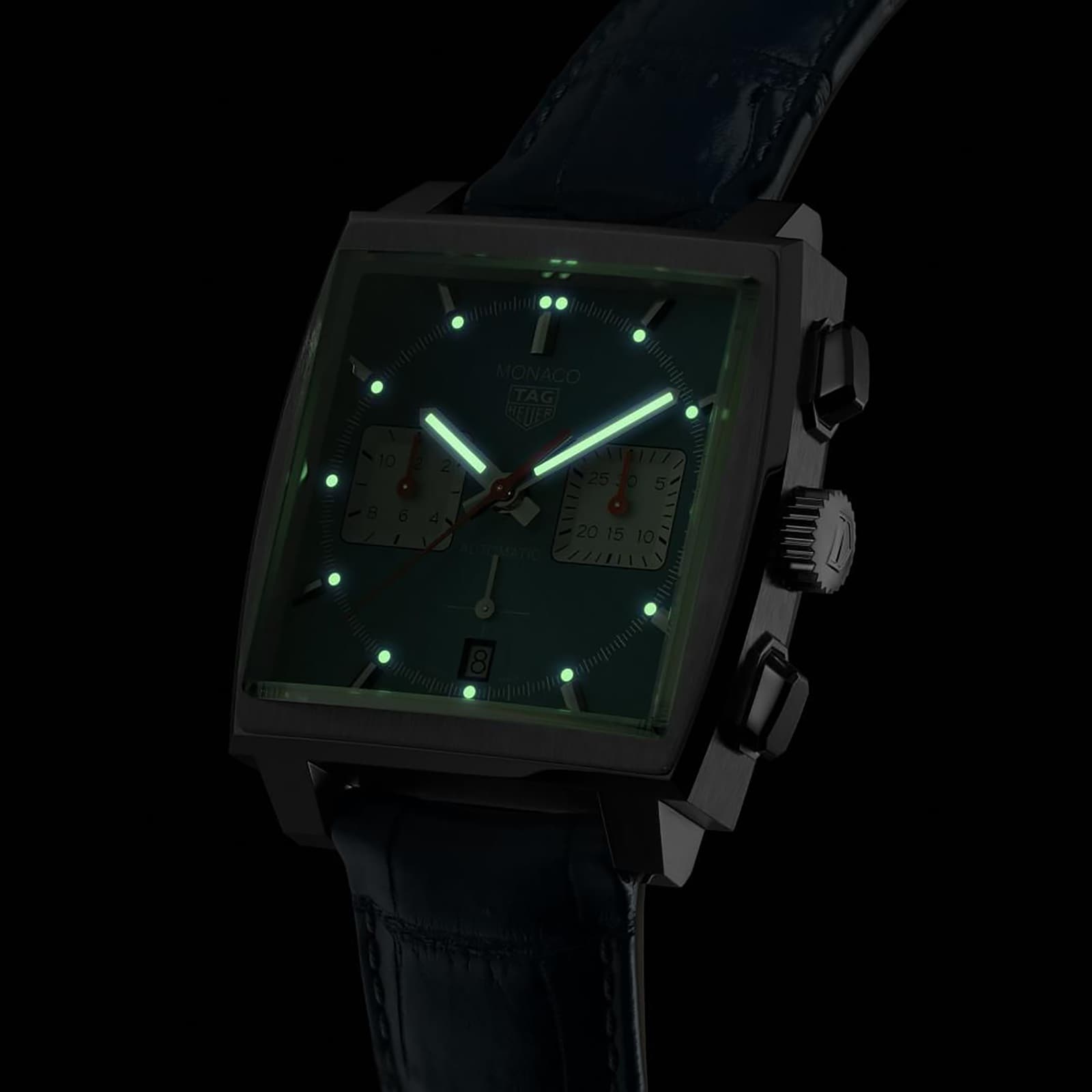 A TAG Heuer Monaco Chronograph watch with glowing green hour markers and hands in a dark setting.
