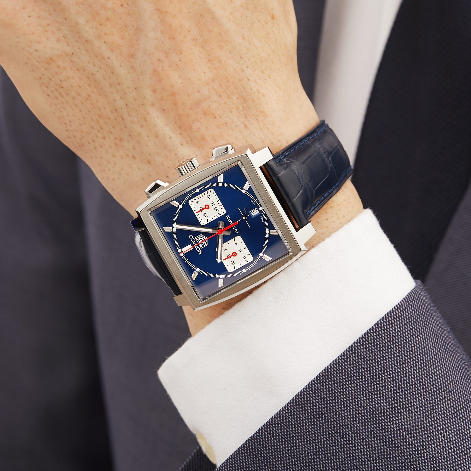 A person in a dark suit and white shirt showcases the TAG Heuer Monaco Chronograph, featuring a square case, blue dial, black leather strap, and Heuer 02 movement.