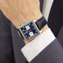 A person in a dark suit and white shirt showcases the TAG Heuer Monaco Chronograph, featuring a square case, blue dial, black leather strap, and Heuer 02 movement.