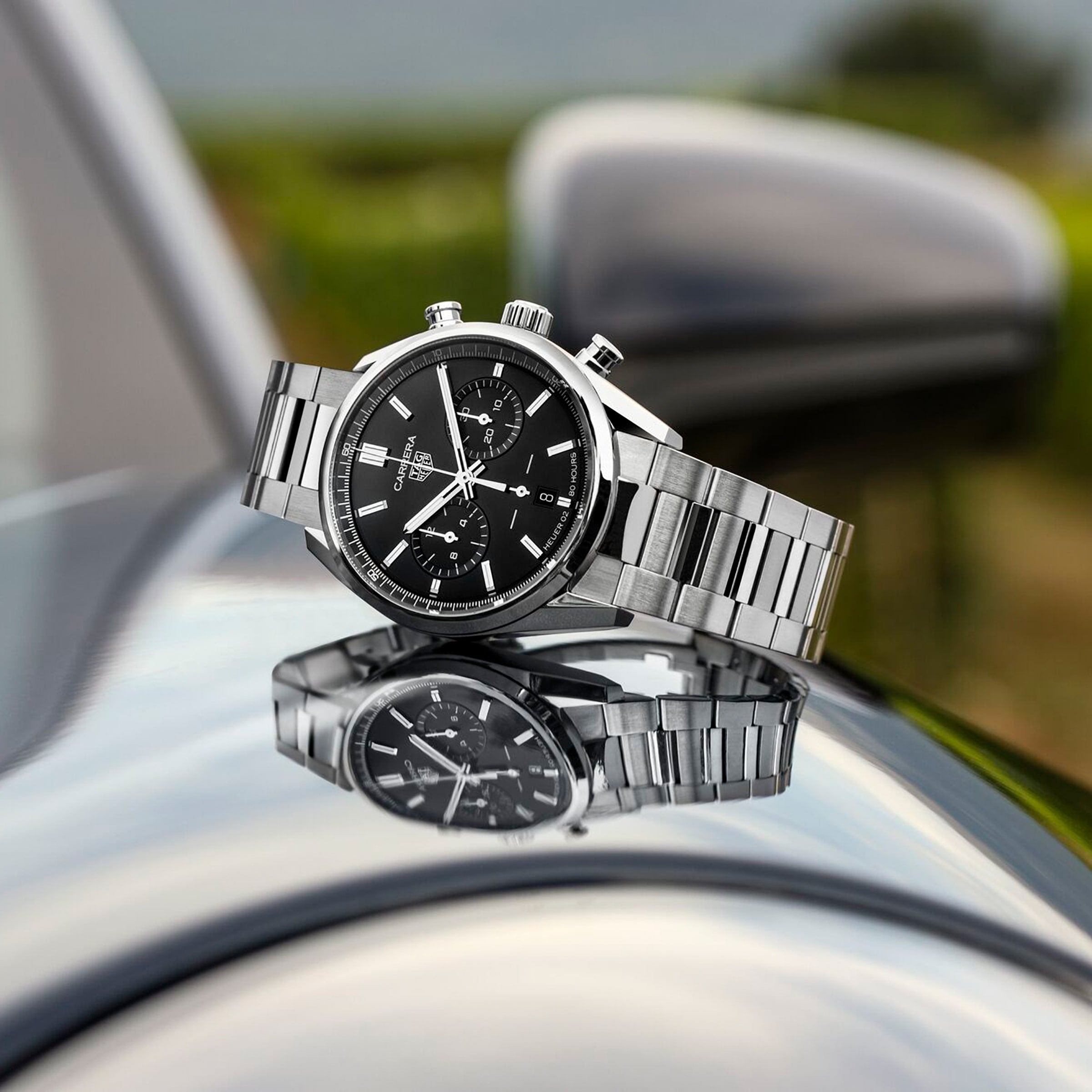 A TAG Heuer Carrera Chronograph wristwatch with a black dial sits atop a metallic car surface, its reflection highlighting the watchs iconic motor-racing heritage.