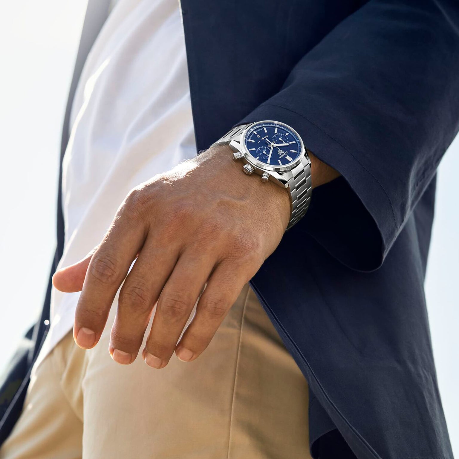 Wearing a navy blazer, white shirt, beige pants, and a TAG Heuer Carrera Chronograph wristwatch with a blue face—an iconic piece inspired by motor-racing heritage.