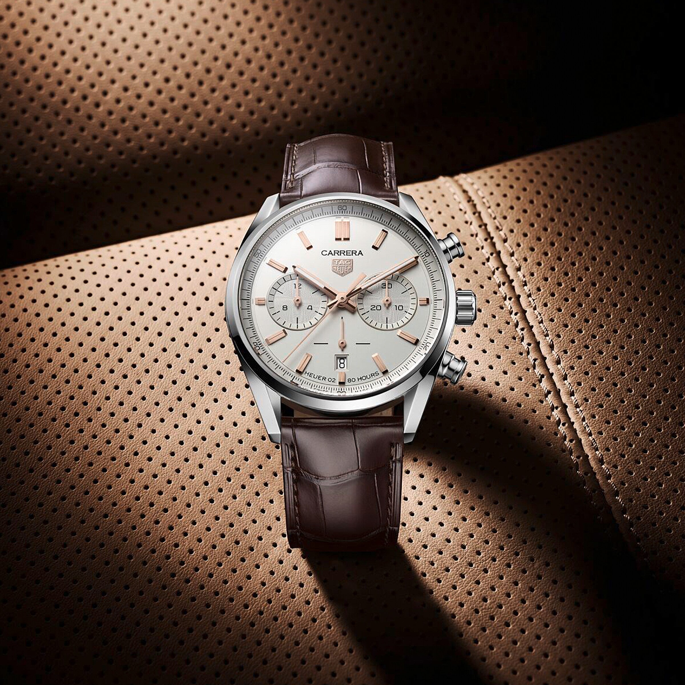A TAG Heuer Carrera Chronograph wristwatch with a silver case and brown leather strap is displayed on perforated tan leather upholstery.