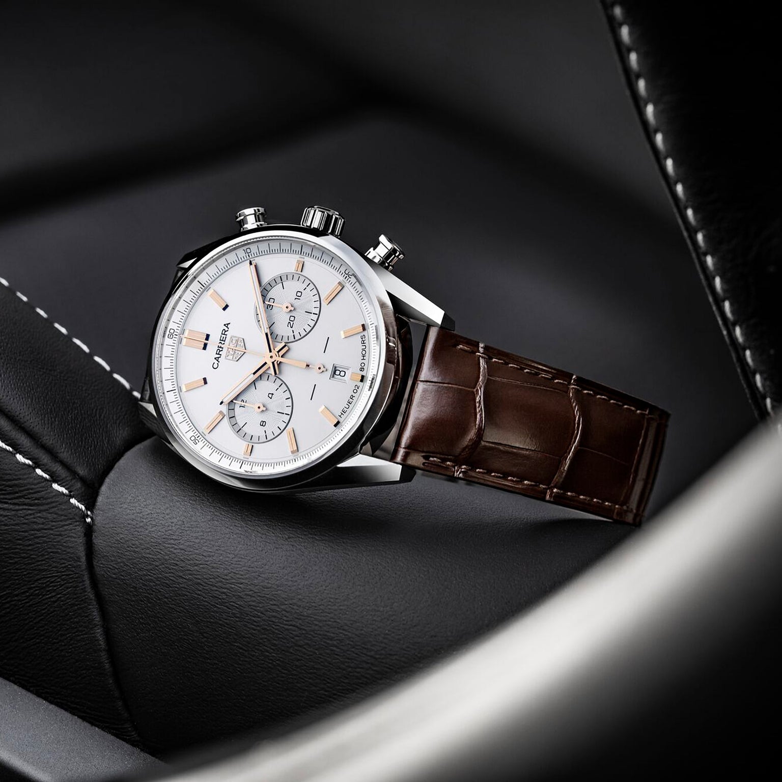 A TAG Heuer Carrera Chronograph with a brown leather strap and white dial rests on a black leather car seat with white stitching, displaying its refined automatic movement.