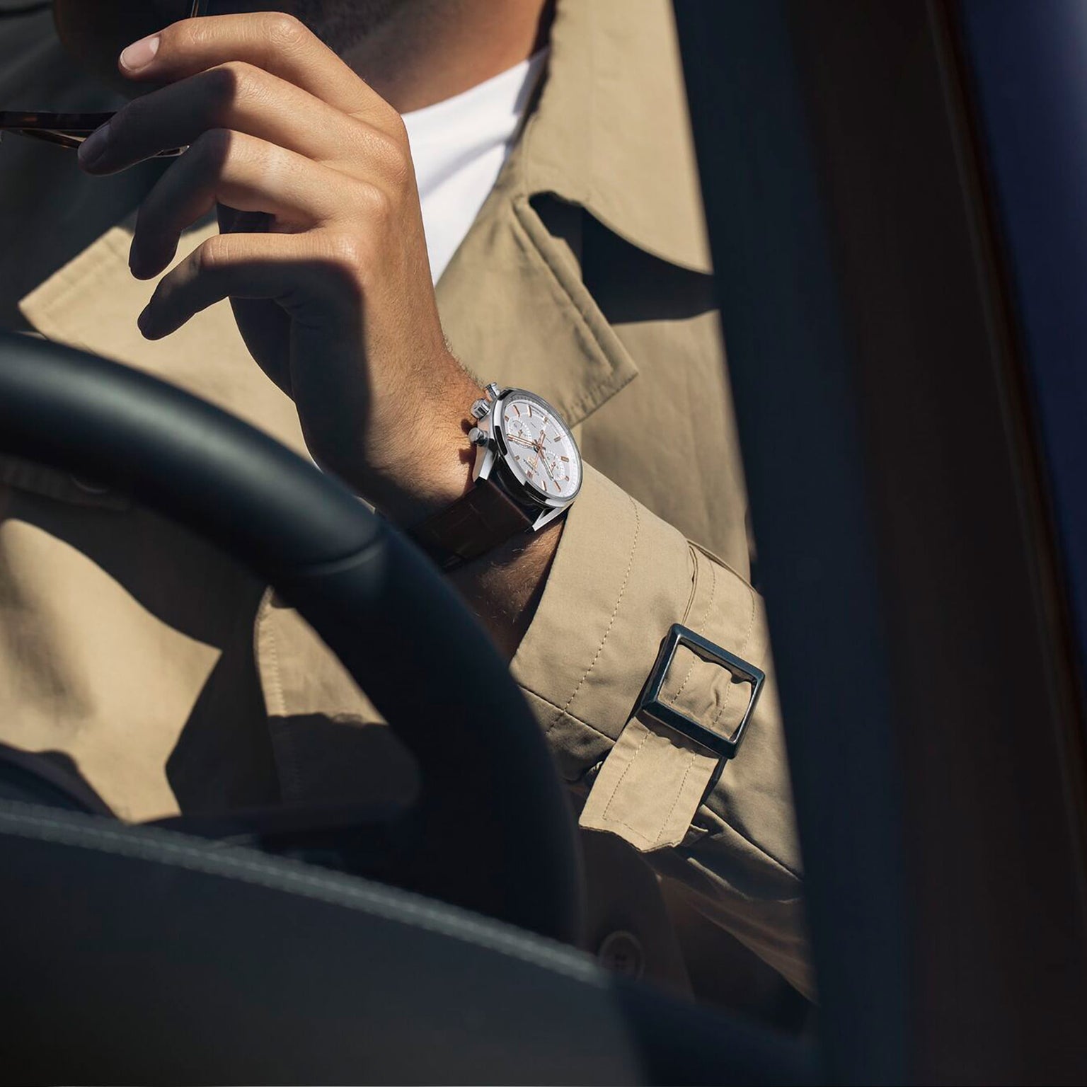A person wearing a beige trench coat and TAG Heuer Carrera Chronograph wristwatch sits in a car, holding Carrera eyeglasses near their face.