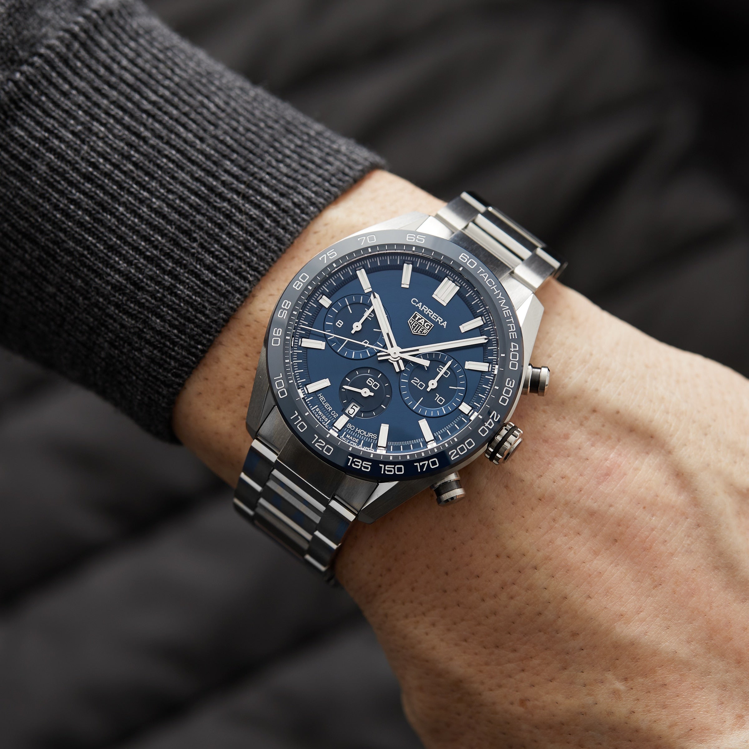 A TAG Heuer Carrera Chronograph is worn on a person’s left wrist, with its stainless steel case and blue dial visible against a gray long-sleeve shirt.