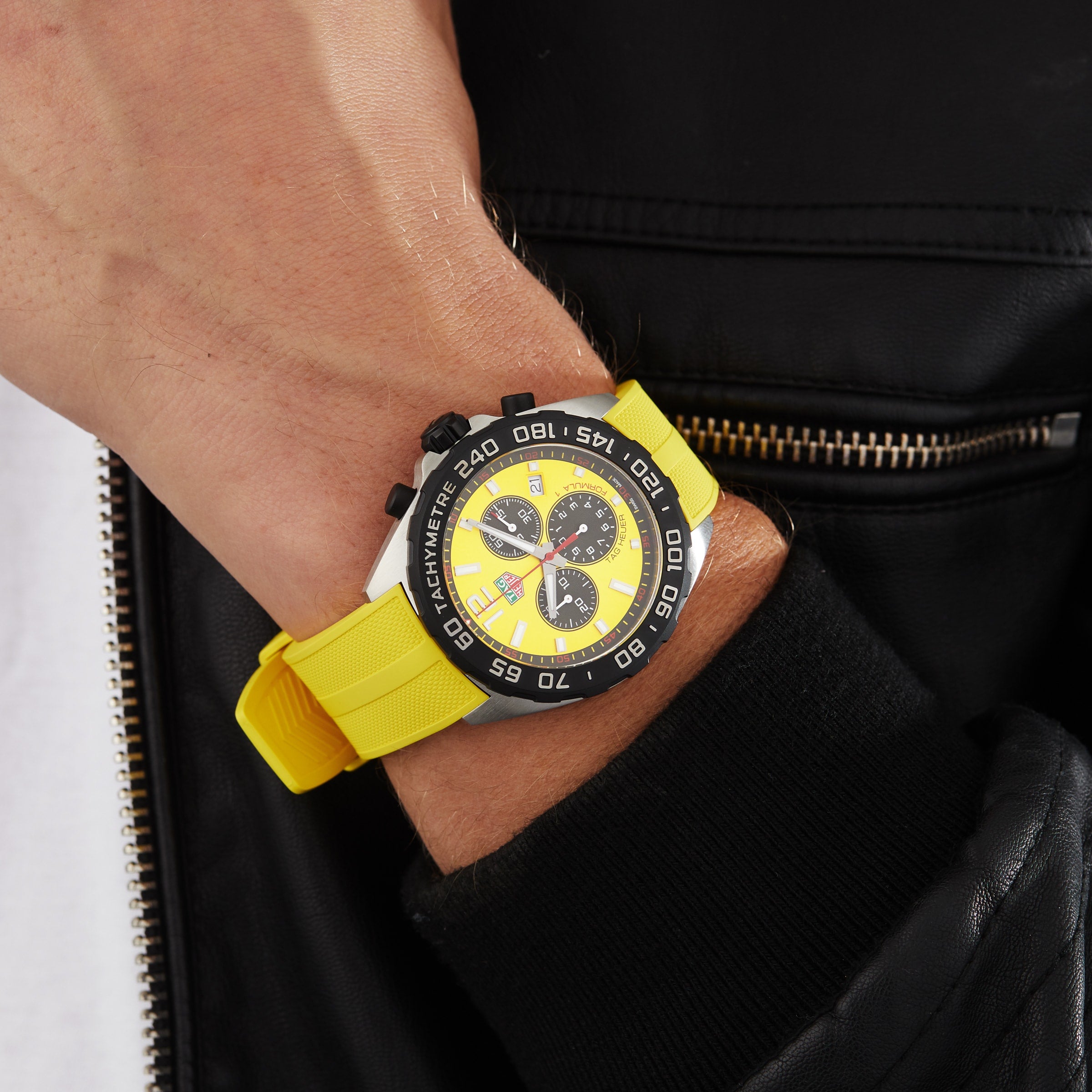 A person wearing a black leather jacket and a yellow TAG Heuer Formula 1 Chronograph wristwatch with a black and yellow face, tachymeter bezel, and precise quartz movement.