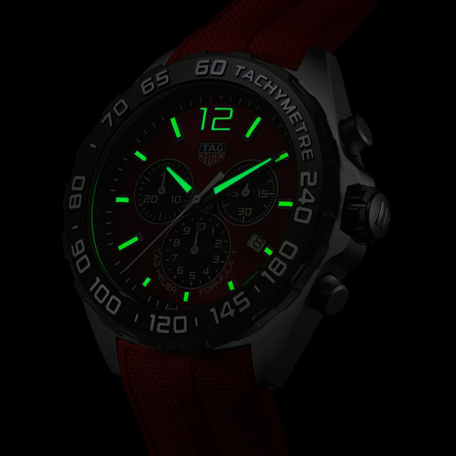 Close-up of a TAG Heuer Formula 1 Chronograph in low light, with green luminescent hands and numerals, stainless steel case, and partially visible red strap.