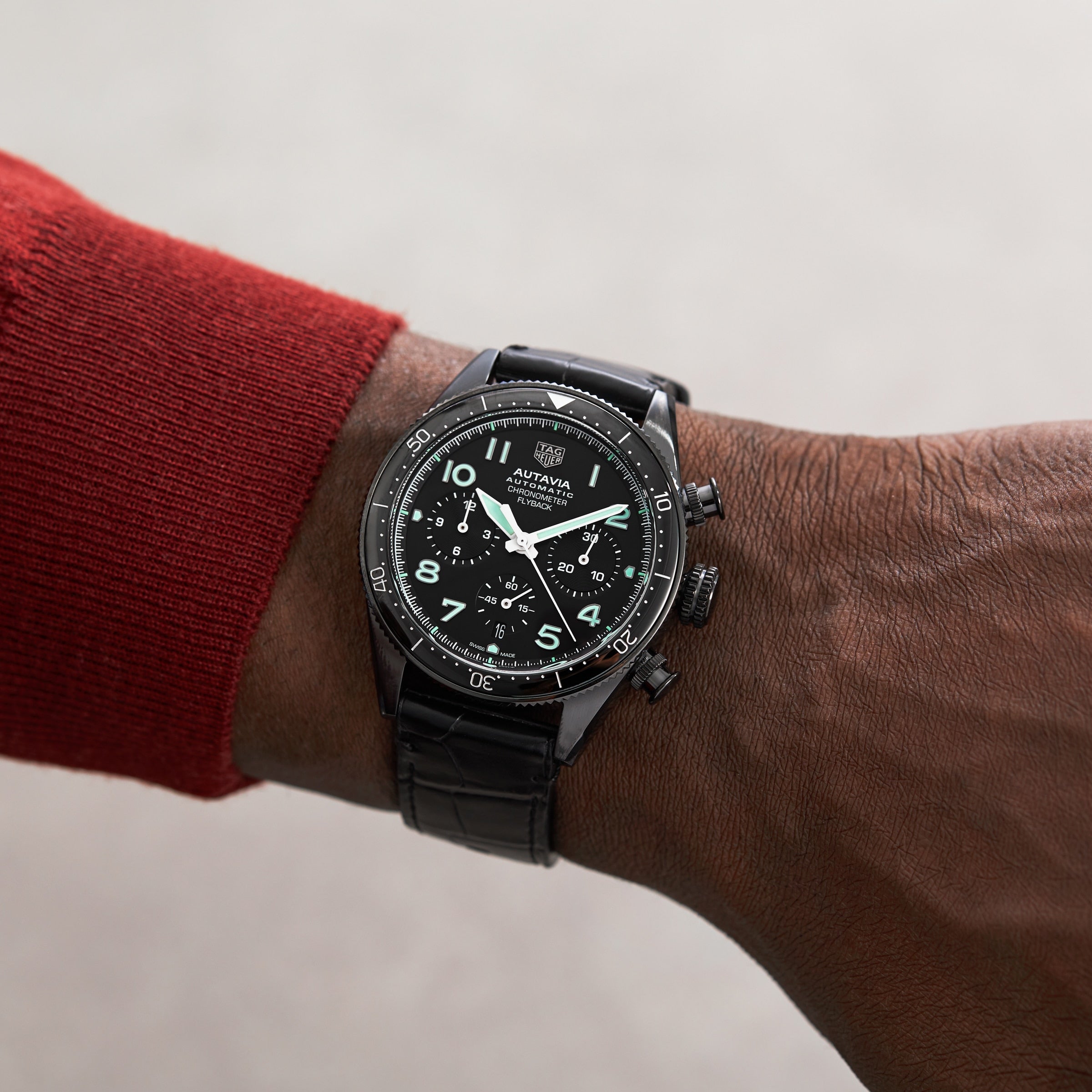 A TAG Heuer Autavia Flyback Chronometer with luminous hands and numerals, featuring a calibre 02 Flyback movement, is worn on the left wrist of a person in a red long-sleeve shirt.
